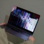 Osoba trzyma laptop Toshiba z pękniętym, kolorowo zakłóconym ekranem