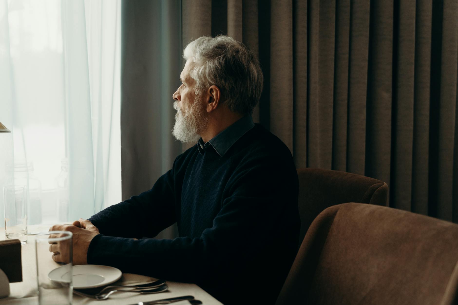 Starszy mężczyzna z brodą zamyślony przy stole, patrzy przez okno